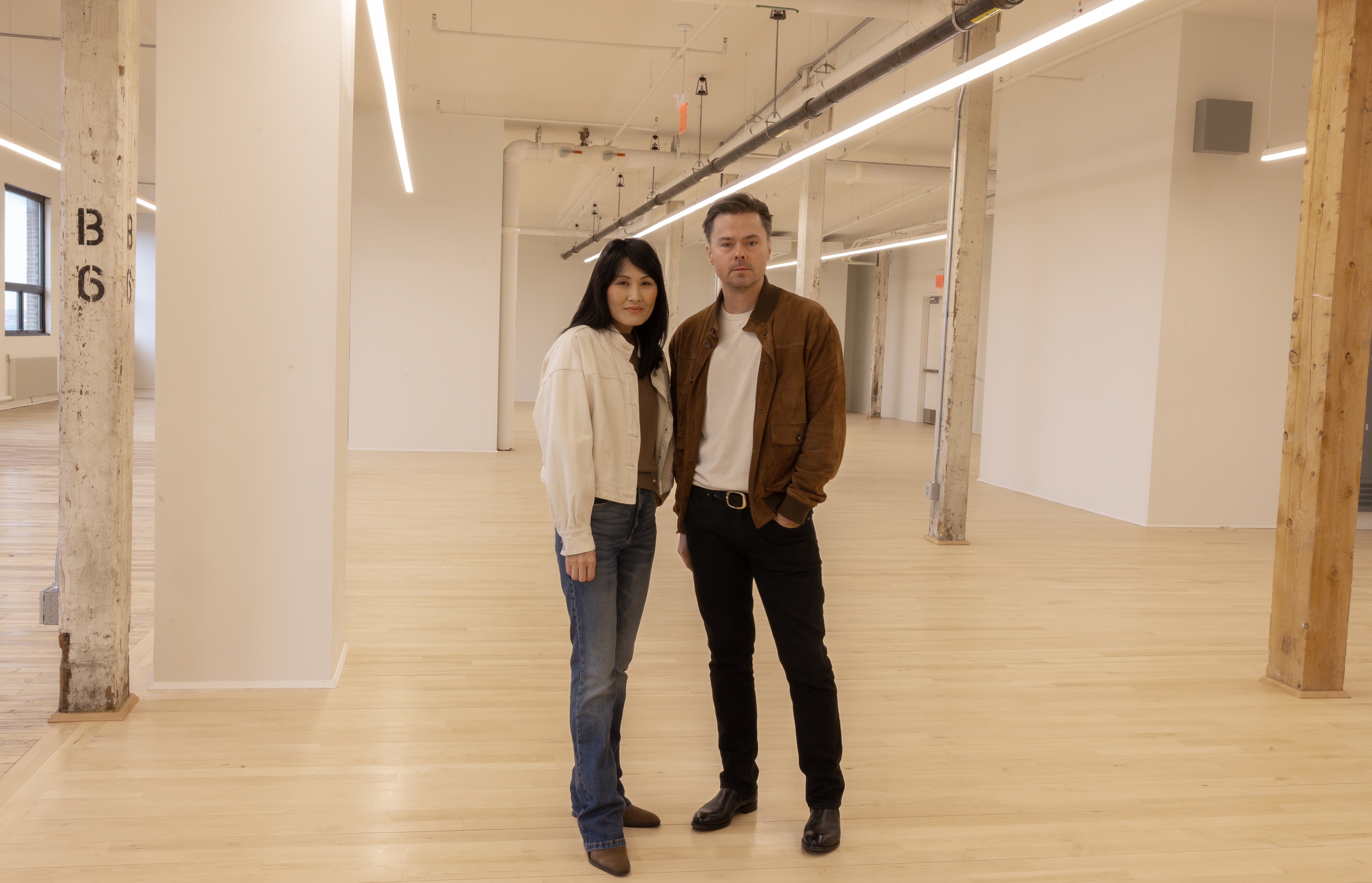 Jean and Oliver Pelle stand side by side in the spacious, modern, expanded PELLE headquarters on the fifth floor of MADE Bush Terminal, with wooden floors and exposed beams. Jean is wearing jeans and a white jacket, and Oliver is wearing black pants and a brown jacket.