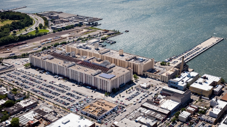 Aerial view of Brooklyn Army Terminal, a large waterfront warehouse complex with numerous parking lots filled with cars. The area is surrounded by industrial buildings, green spaces, and docks extending into the water. Aerial view of Brooklyn Army Terminal, a large waterfront warehouse complex with numerous parking lots filled with cars. The area is surrounded by industrial buildings, green spaces, and docks extending into the water.