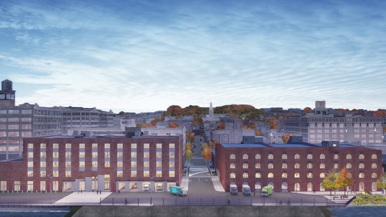 MADE Bush Terminal Campus rendering by nARCHITECTS. A wide view of two adjacent brick buildings with multiple windows set against a New York City cityscape backdrop under a blue sky with clouds. A few vehicles are parked along the street.