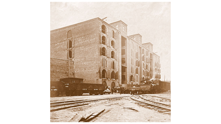 Sepia-toned photo of a large brick warehouse with arched windows and several floors. Railway tracks and train cars sit in front, with people visible near the building’s entrance. Sepia-toned photo of a large brick warehouse with arched windows and several floors. Railway tracks and train cars sit in front, with people visible near the building’s entrance.