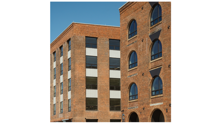 Two adjacent brick buildings with modern windows. One building has rectangular windows in vertical rows, while the other features arched windows with black triangular accents. The sky is clear and blue.