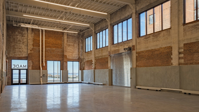 Spacious industrial warehouse with high ceilings, exposed brick walls, large windows letting in natural light, and a rolled-up metal garage door; outside, water and buildings are visible through the windows and glass doors.