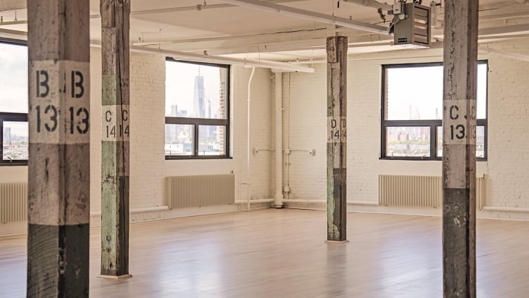 Spacious industrial loft with wooden pillars labeled B13 and C14, large windows offering city skyline views, and light wood flooring. Walls are white brick with a clean, minimalist appearance.
