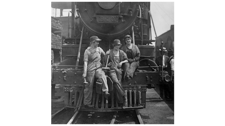 Three female railroad workers in work clothes and hats sit on the front of a large steam locomotive, smiling. Three female railroad workers in work clothes and hats sit on the front of a large steam locomotive, smiling.