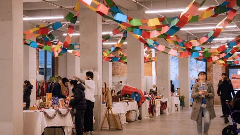 A spacious indoor market with colorful paper garlands hanging from the ceiling. Several people browse tables displaying various items like clothes, art, and crafts. A woman walks past with a stroller on the right.