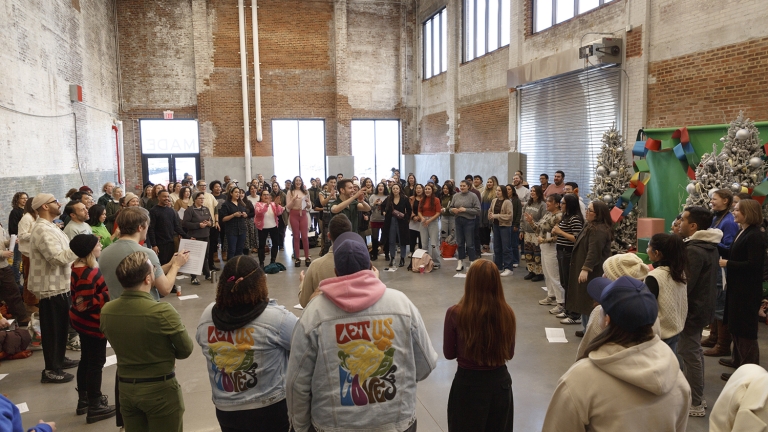 Show thumbnail preview A large, diverse group of people stands in a spacious, industrial-style room with brick walls and high windows, gathered in a circle around performers wearing colorful Love Us Love jackets, near decorated Christmas trees.