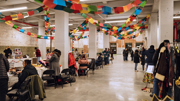 A spacious indoor market with people browsing vendor tables. Colorful paper garlands hang from the ceiling, and clothing racks and crafts are on display. The atmosphere is lively and festive.