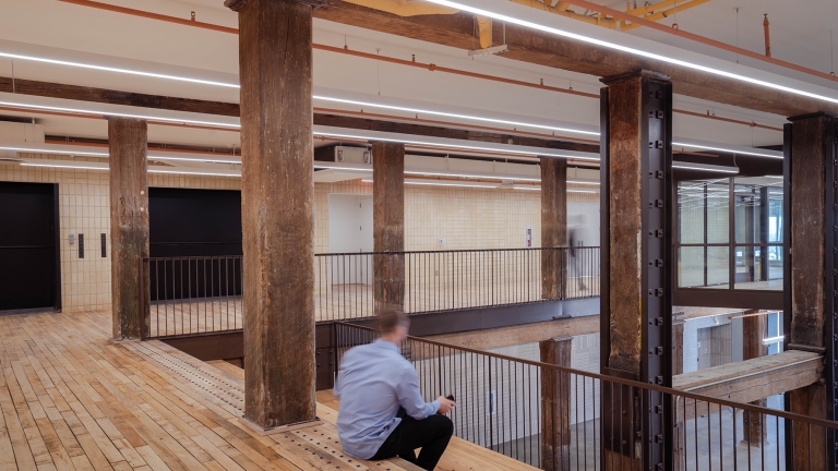 A person sits on a wooden bench in an industrial-style interior with exposed beams, wooden floors, metal railings, and cream tile walls, creating a spacious, modern atmosphere.