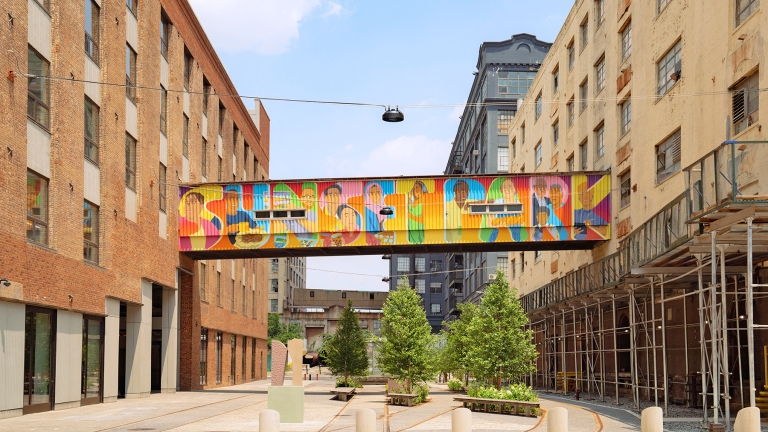A colorful pedestrian bridge with the words SUNSET PARK connects two industrial buildings over a walkway lined with young trees and modern art sculptures. The sky is partly cloudy.