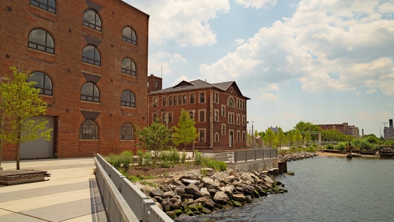 Red brick industrial buildings sit beside a river, surrounded by trees and rocks along the water’s edge. The sky is partly cloudy, and the area appears calm and well-maintained.