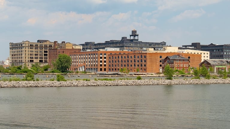View across a calm river toward a row of brick and industrial buildings, with trees and greenery along the riverside and a partly cloudy sky above. Leaves partially frame the scene at the top of the image.