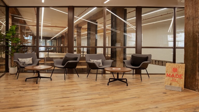 A modern office lounge with four gray armchairs, two round wooden tables, a plant, and a MADE sign on the wooden floor. Large windows with wooden beams separate the lounge from another room.