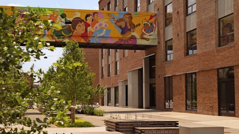 A modern brick building with large windows and a colorful mural on a skybridge above a courtyard, featuring wooden benches, young trees, and a clear, sunny sky.