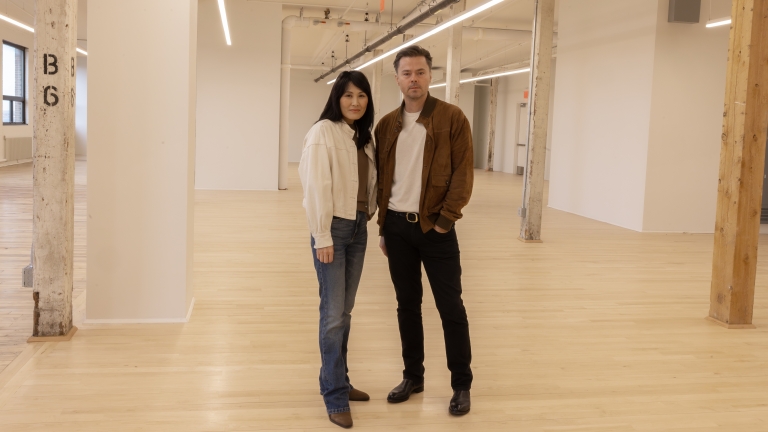 Jean and Oliver Pelle stand side by side in the spacious, modern, expanded PELLE headquarters on the fifth floor of MADE Bush Terminal, with wooden floors and exposed beams. Jean is wearing jeans and a white jacket, and Oliver is wearing black pants and a brown jacket.