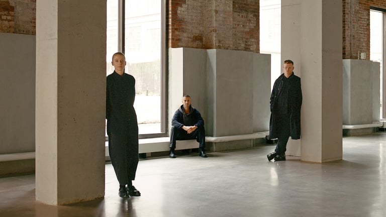 Three people in dark clothing pose separately in a spacious, industrial-style room with large windows, exposed brick walls, and concrete pillars; two stand near pillars and one sits on a windowsill.