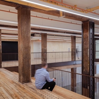 A person sits on a wooden bench in an industrial-style interior with exposed beams, wooden floors, metal railings, and cream tile walls, creating a spacious, modern atmosphere. A person sits on a wooden bench in an industrial-style interior with exposed beams, wooden floors, metal railings, and cream tile walls, creating a spacious, modern atmosphere.