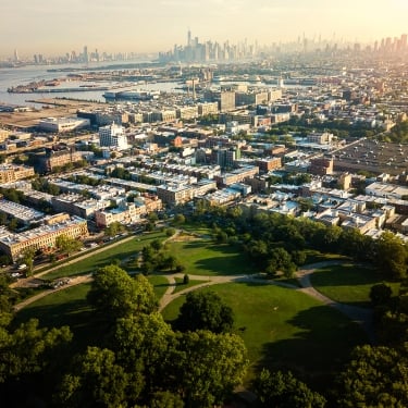 Aerial view of a large city park with green lawns and trees in the foreground, surrounded by dense urban buildings and a distant skyline under a hazy sky. Aerial view of a large city park with green lawns and trees in the foreground, surrounded by dense urban buildings and a distant skyline under a hazy sky.