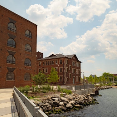 Red brick industrial buildings sit beside a river, surrounded by trees and rocks along the water’s edge. The sky is partly cloudy, and the area appears calm and well-maintained. Red brick industrial buildings sit beside a river, surrounded by trees and rocks along the water’s edge. The sky is partly cloudy, and the area appears calm and well-maintained.