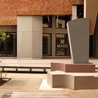 A modern outdoor courtyard with abstract concrete benches and a geometric sculpture, set against a brick building with large windows. One window displays the words MADE in bold letters. A modern outdoor courtyard with abstract concrete benches and a geometric sculpture, set against a brick building with large windows. One window displays the words MADE in bold letters.
