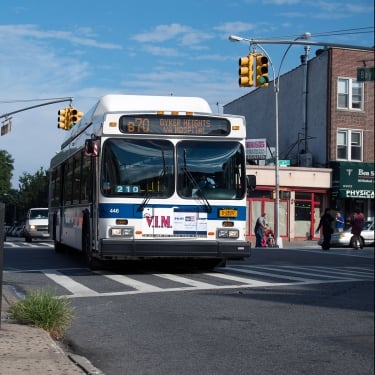 A city bus labeled B70 to Dyker Heights approaches a crosswalk at an intersection on a sunny day, with buildings, traffic lights, and pedestrians in the background. A city bus labeled B70 to Dyker Heights approaches a crosswalk at an intersection on a sunny day, with buildings, traffic lights, and pedestrians in the background.