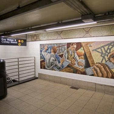 Mural in a subway station shows two men at a desk examining maps and architectural plans. Hands and tools are visible. Above, a sign indicates directions to Bay Ridge and Coney Island. The area is lit with overhead fluorescent lights. Mural in a subway station shows two men at a desk examining maps and architectural plans. Hands and tools are visible. Above, a sign indicates directions to Bay Ridge and Coney Island. The area is lit with overhead fluorescent lights.