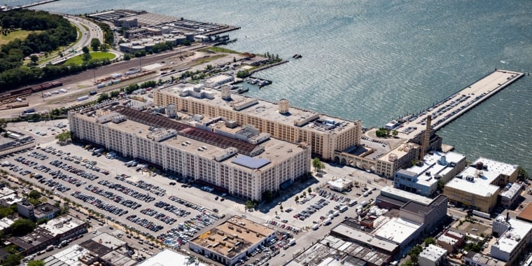 Aerial view of Brooklyn Army Terminal, a large waterfront warehouse complex with numerous parking lots filled with cars. The area is surrounded by industrial buildings, green spaces, and docks extending into the water.