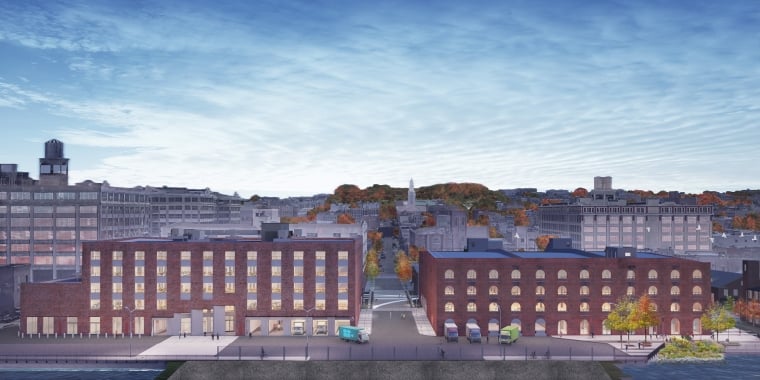 MADE Bush Terminal Campus rendering by nARCHITECTS. A wide view of two adjacent brick buildings with multiple windows set against a New York City cityscape backdrop under a blue sky with clouds. A few vehicles are parked along the street.