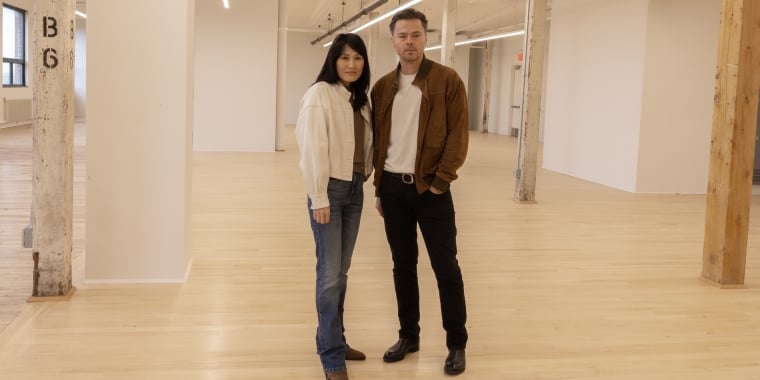 Jean and Oliver Pelle stand side by side in the spacious, modern, expanded PELLE headquarters on the fifth floor of MADE Bush Terminal, with wooden floors and exposed beams. Jean is wearing jeans and a white jacket, and Oliver is wearing black pants and a brown jacket.