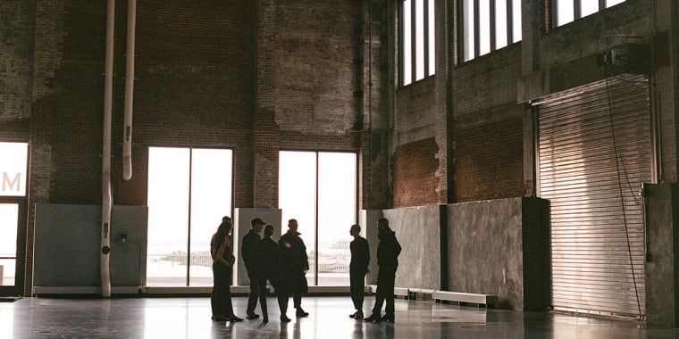 A group of people stand and talk inside a large, industrial warehouse with tall windows, exposed brick walls, and a high ceiling. Light streams in through the windows, casting shadows on the floor.