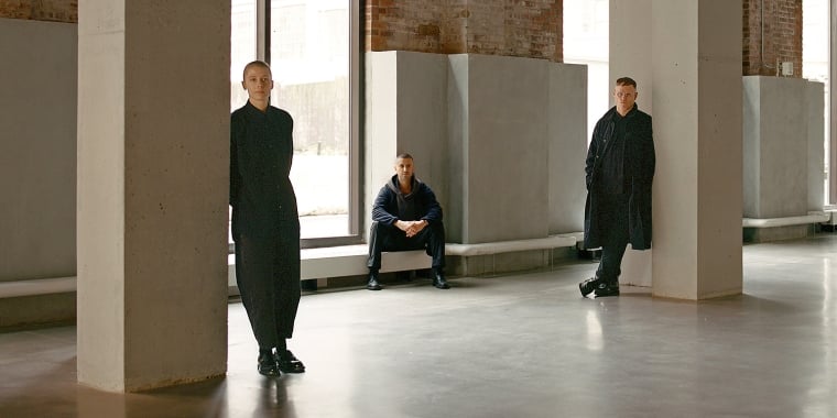 Three people in dark clothing pose separately in a spacious, industrial-style room with large windows, exposed brick walls, and concrete pillars; two stand near pillars and one sits on a windowsill.