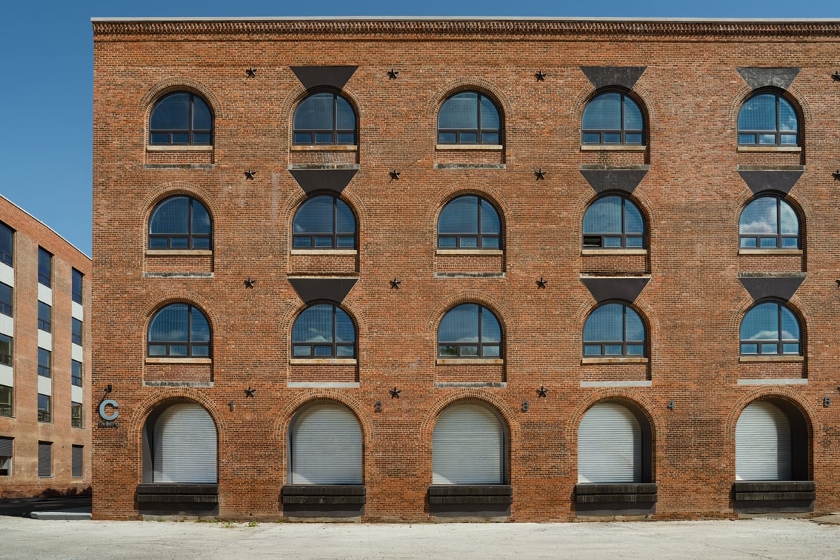 A brick building with three floors and three arched windows on each level, each window framed with dark accents. The ground floor windows appear to have closed metal shutters. The sky is clear and blue. A brick building with three floors and three arched windows on each level, each window framed with dark accents. The ground floor windows appear to have closed metal shutters. The sky is clear and blue.