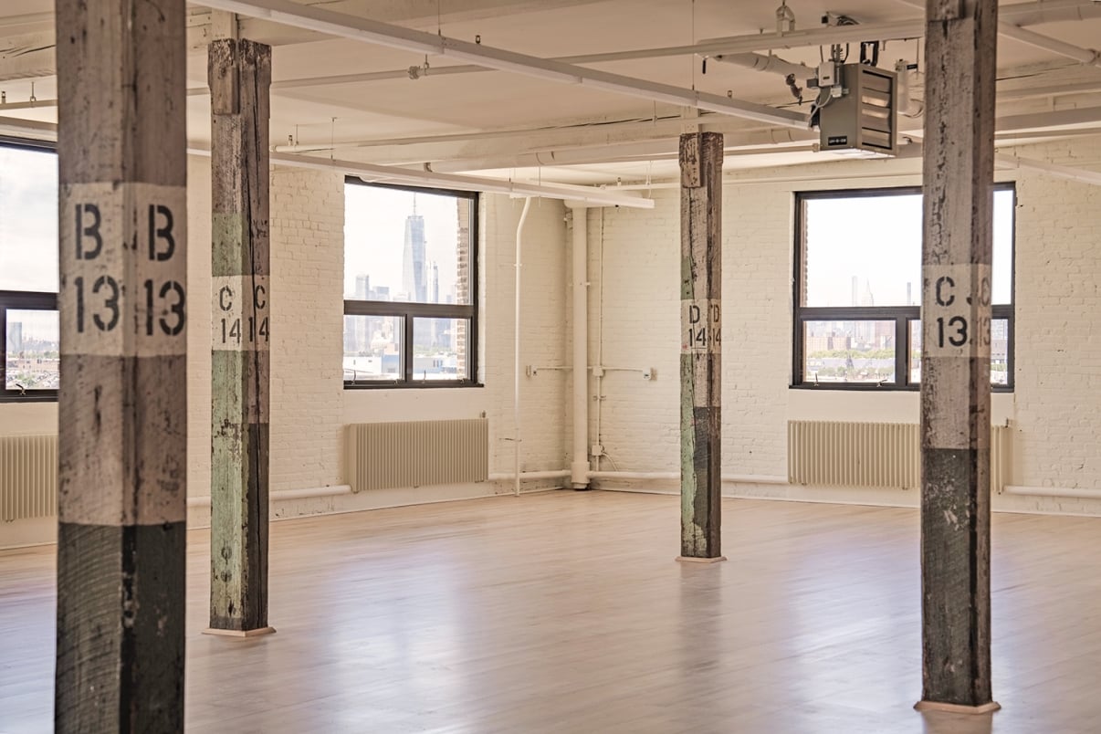 Spacious industrial loft with wooden pillars labeled B13 and C14, large windows offering city skyline views, and light wood flooring. Walls are white brick with a clean, minimalist appearance.