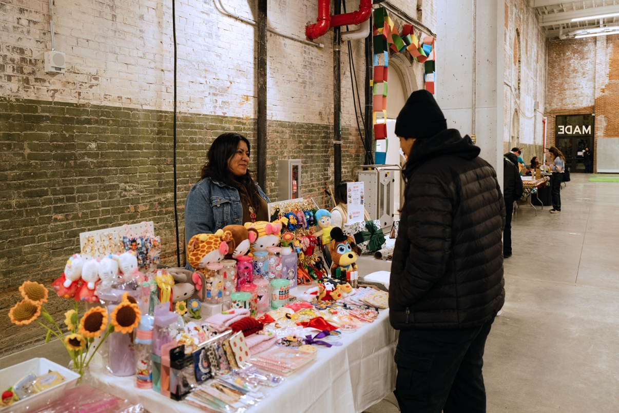 Show thumbnail preview A vendor smiles at a customer at an indoor market booth displaying colorful handmade crafts, plush toys, flowers, and stationery, with a brick wall and decorative archway in the background.
