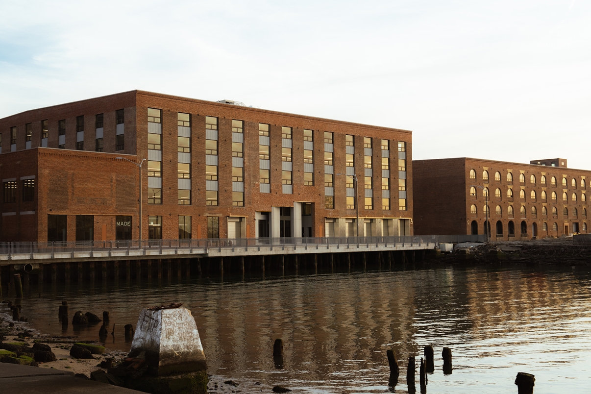 A large, rectangular brick building with many windows stands beside a calm waterfront, reflecting in the water. Nearby, weathered wooden posts emerge from the shoreline. The scene is peaceful and bathed in soft light.