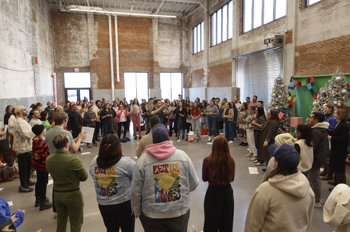 Show thumbnail preview A large, diverse group of people stands in a spacious, industrial-style room with brick walls and high windows, gathered in a circle around performers wearing colorful Love Us Love jackets, near decorated Christmas trees.