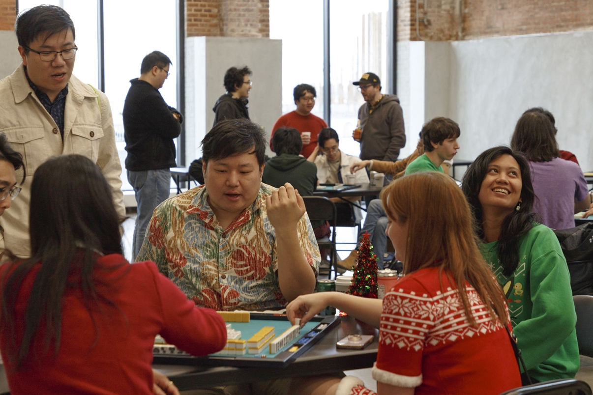 Show thumbnail preview A group of people sits around tables playing games indoors. In the foreground, four people play a board game while others socialize in the background. The atmosphere appears lively and friendly.