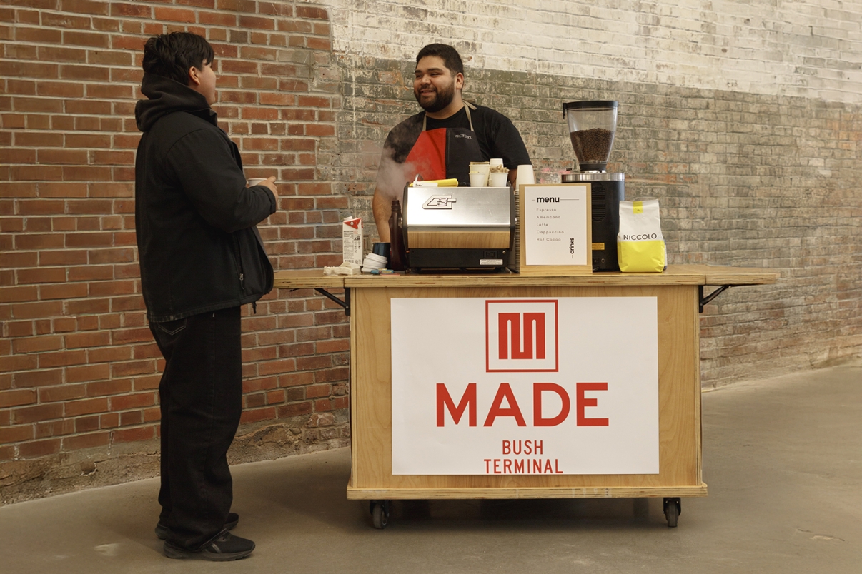 A barista smiles and talks with a customer at a small coffee cart labeled MADE BUSH TERMINAL, set against a brick wall. The cart holds a coffee machine, cups, and coffee supplies.