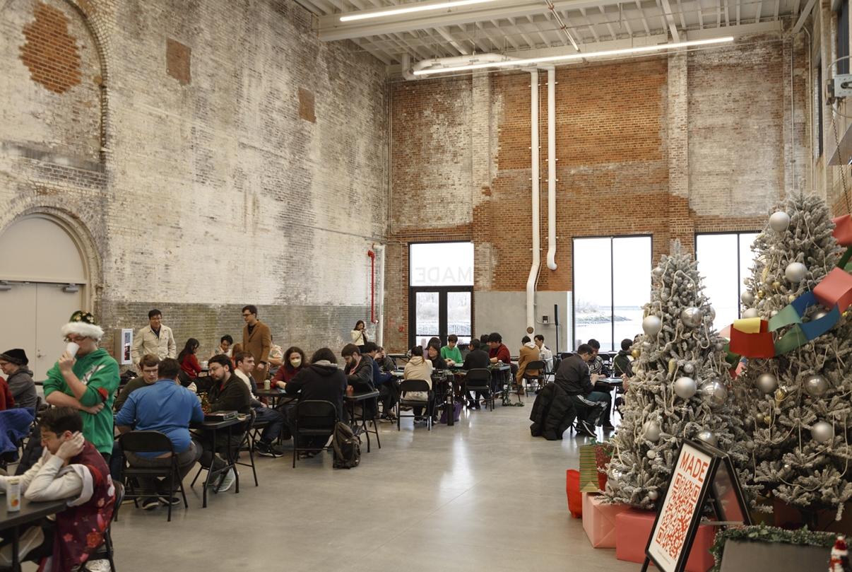 People sit at tables in a spacious, industrial-style room with tall brick walls and large windows. On the right, decorated white Christmas trees and colorful presents are displayed, adding a festive atmosphere.