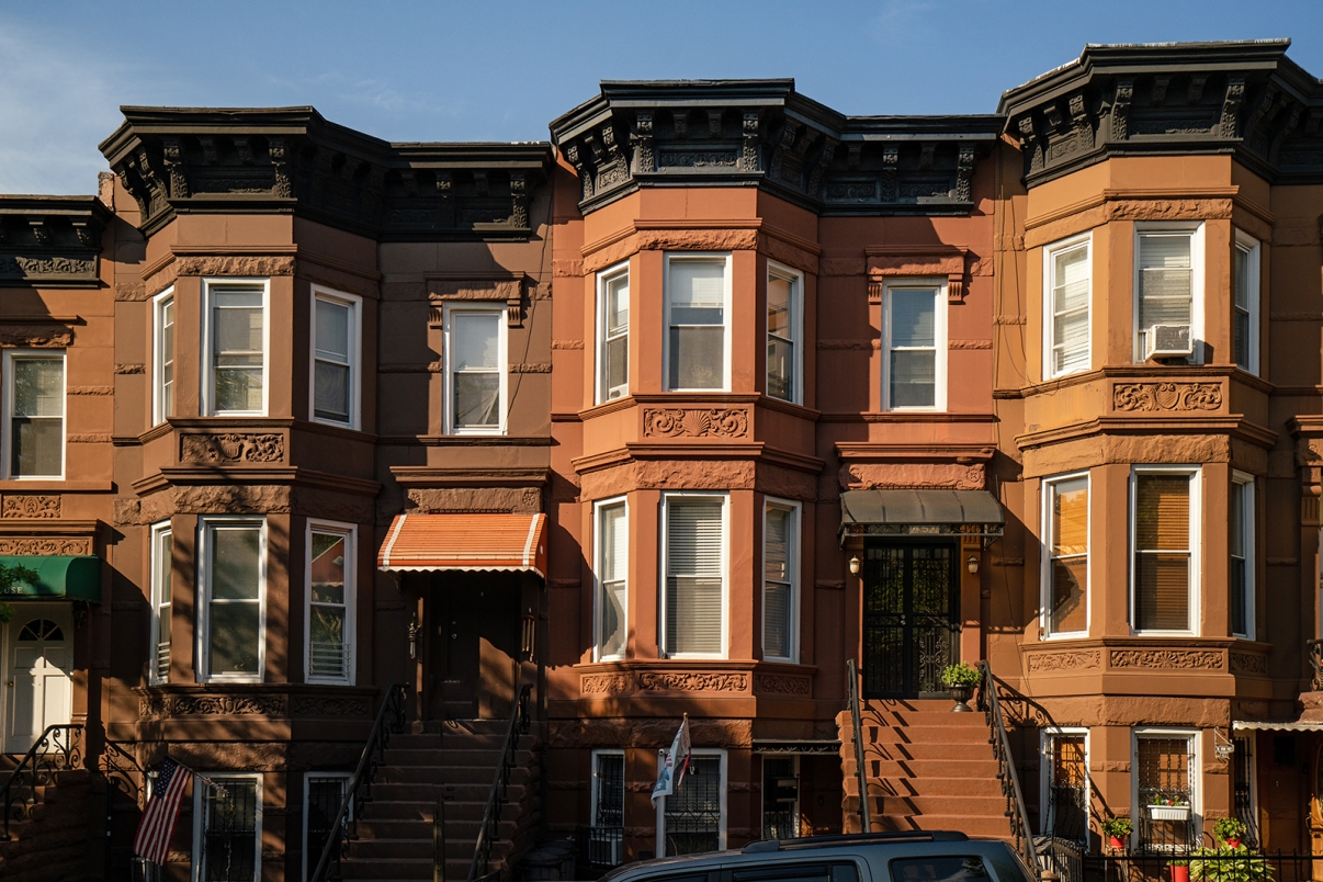 A row of brownstone townhouses with ornate details, stoops leading up to the entrances, and large bay windows. One has an orange awning above the door. A parked car is partially visible in front.