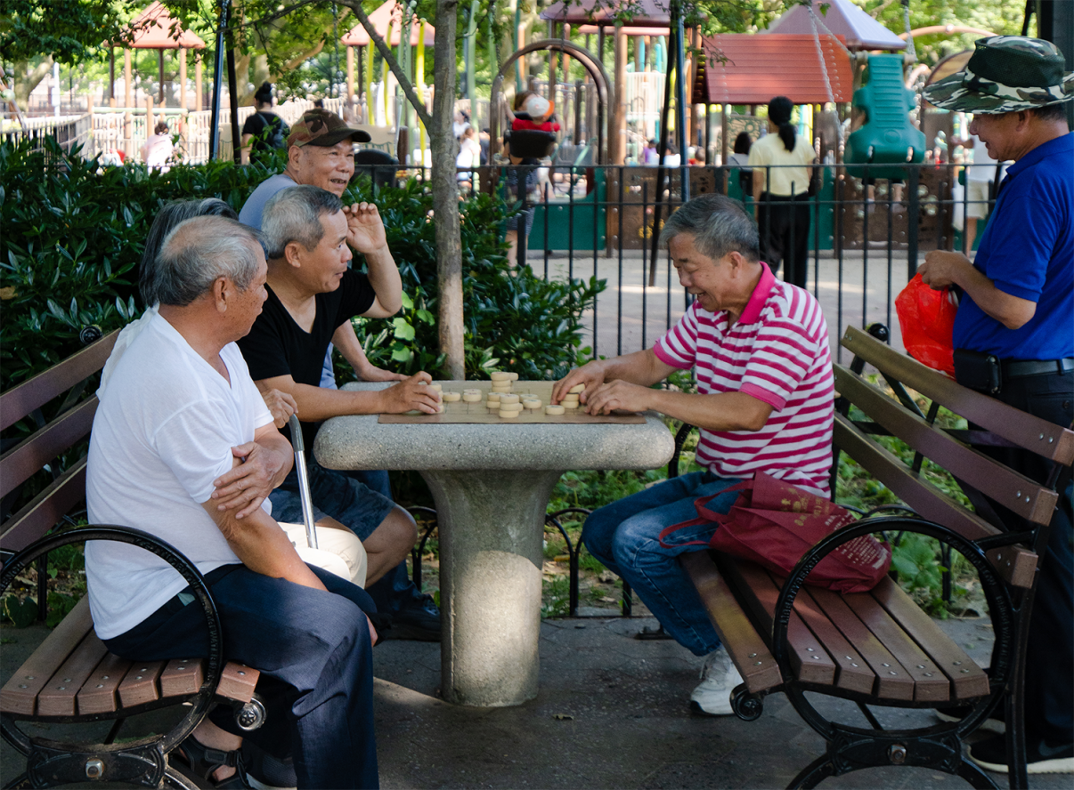 Five elderly men sit and stand around a stone table playing Banqi in Sunset Park, playing a game. Two benches face the table, trees and greenery surround them, and a playground is visible in the background. Sunlight filters through the leaves.