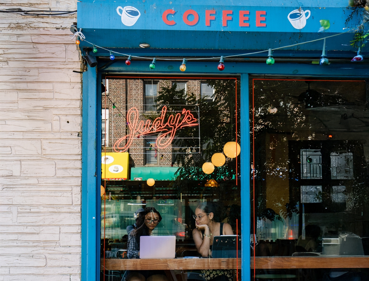 Two people sit at a table with laptops inside a café called Judy’s, seen through a large window. Neon and string lights decorate the window, and a sign above reads COFFEE with coffee cup illustrations.