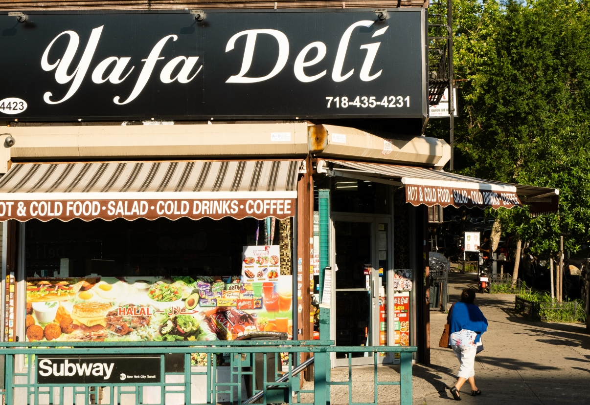 A person walks past Yafa Deli, a corner store with a black sign and awnings, located near a subway entrance. Bright morning sunlight casts shadows, and green trees are visible in the background.