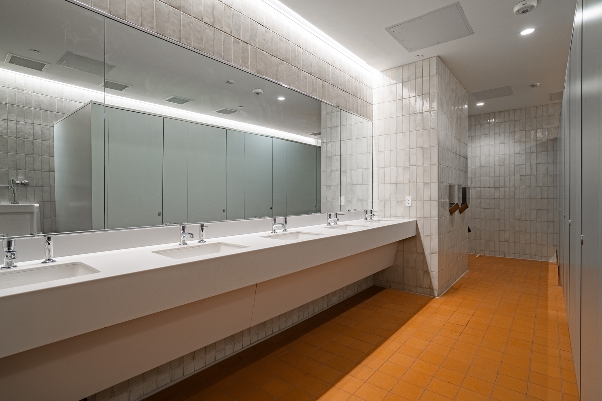 Modern public restroom with a long white sink counter, multiple faucets, large wall mirror, tiled walls, orange-brown floor tiles, and gray bathroom stalls. The space is brightly lit and clean.