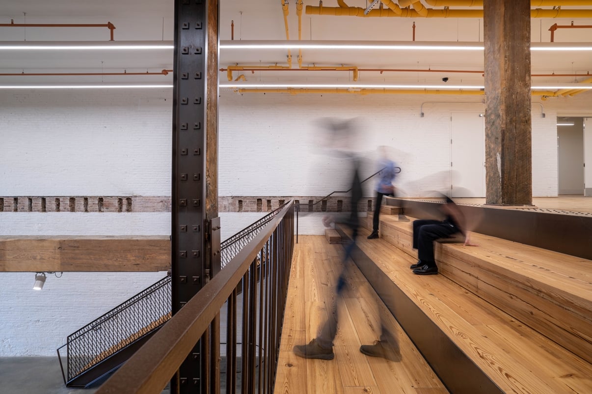 A modern interior with wooden stadium-style seating and exposed beams; three blurred figures move across and up the steps, suggesting motion. White brick walls and yellow overhead pipes are visible in the background.