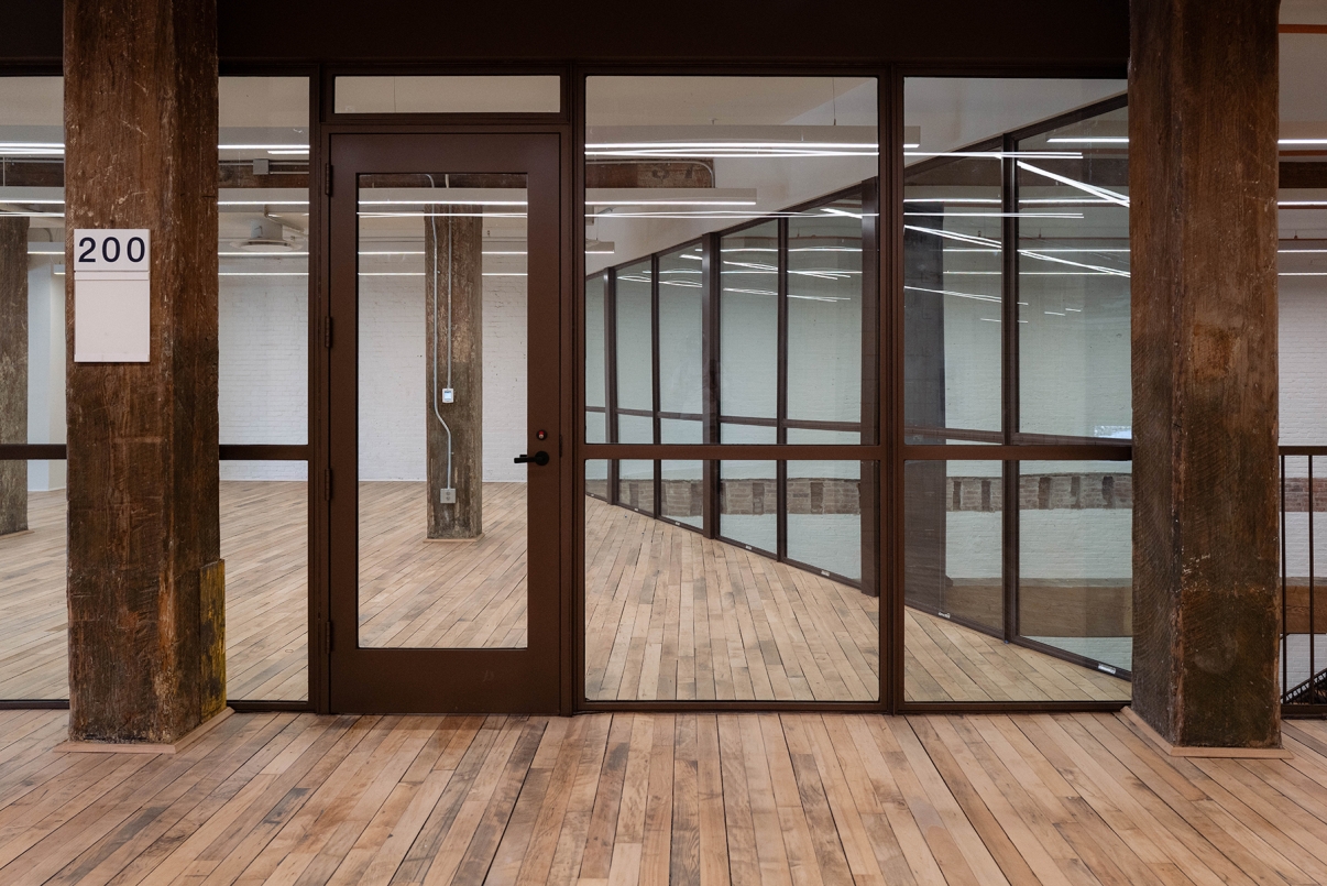 A modern, empty office space with floor-to-ceiling glass walls, a glass door, exposed concrete pillars, and light wood flooring. A sign outside the office displays the number 200. The room is well-lit with ceiling lights.
