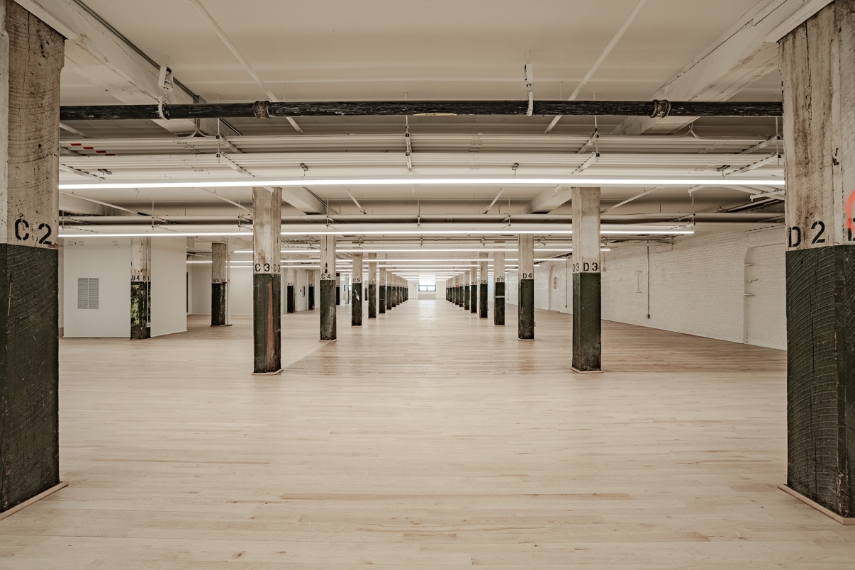A spacious, empty industrial-style interior with light wooden floors, exposed concrete columns, and white walls. Bright overhead lights and a distant window create a sense of depth in the long, open space.