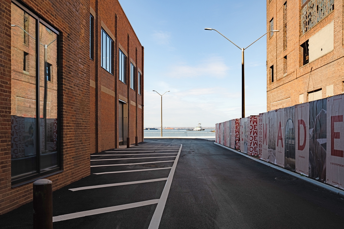 A paved alleyway lined with brick buildings leads toward a body of water, with a distant boat visible. The sky is clear, and a tall fence with red text runs along the right side of the alley.