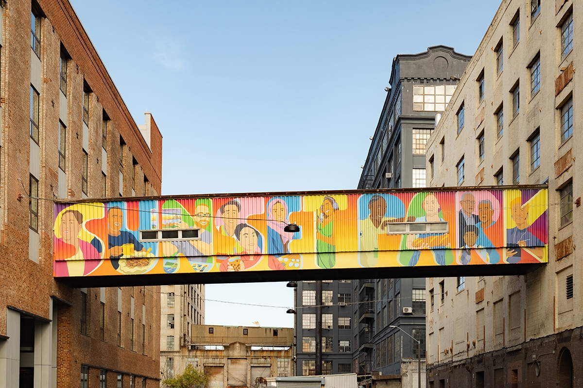 A colorful mural spelling “SUNSET PARK” decorates a skybridge connecting two industrial buildings, with illustrations of people integrated into the vibrant letters.