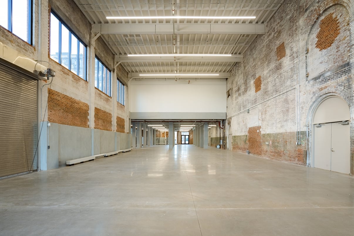 Show thumbnail preview A spacious, empty industrial warehouse with high ceilings, exposed brick and concrete walls, large windows, and polished concrete floors. Light streams in through the windows, creating a bright, open atmosphere.