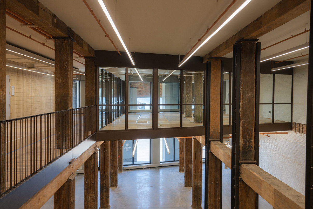 Interior of a modern industrial building with exposed wooden columns, large windows, white brick walls, and a suspended glass walkway connecting upper levels. The space has high ceilings and minimalistic lighting.